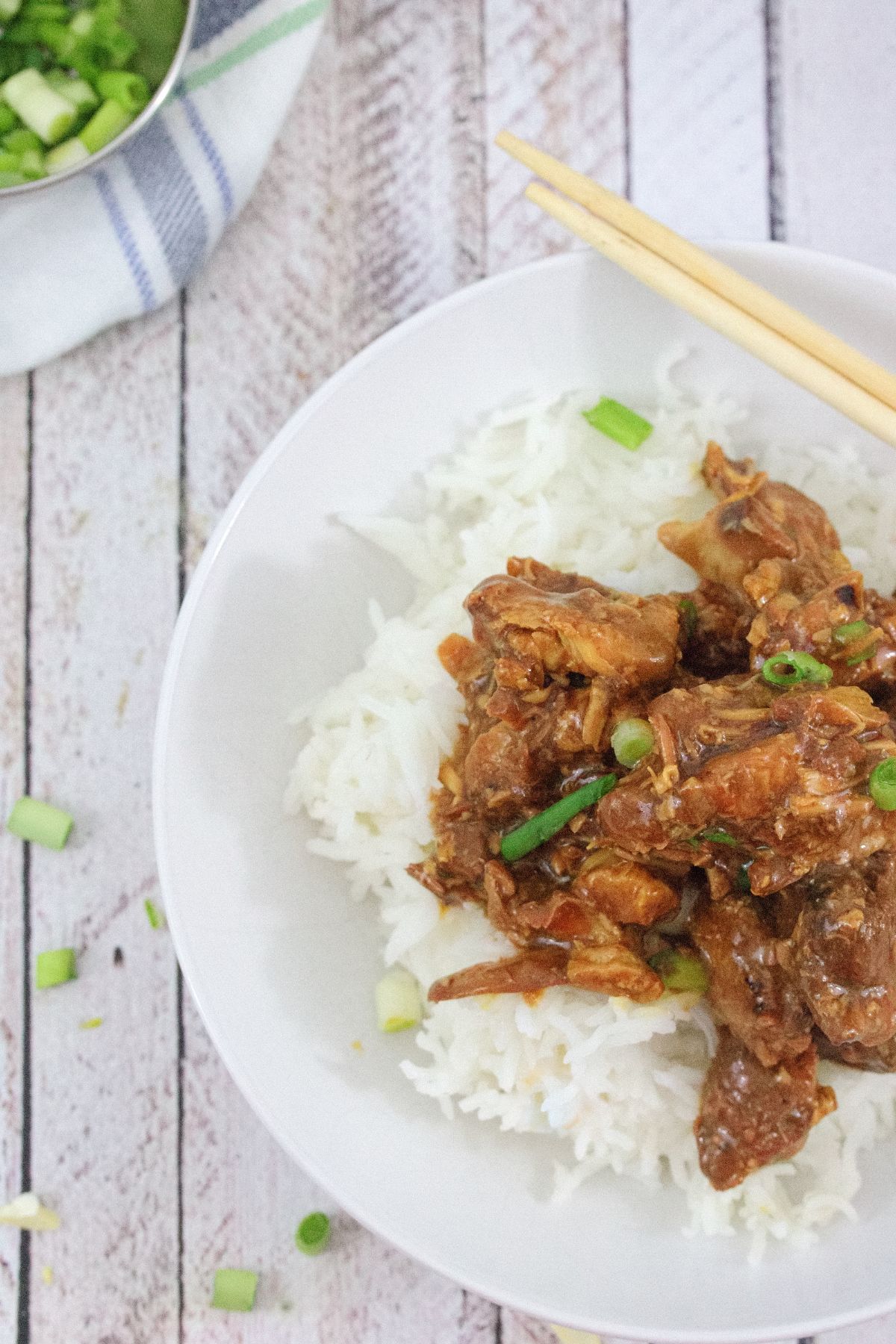 White bowl with rice and teriyaki chicken topped with green onions with chop sticks on the bowl and more green onions behind it.