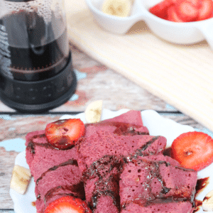 three pink vegan crepes on a white plate with chocolate, strawberries and banana on them and coffee in a french press behind them