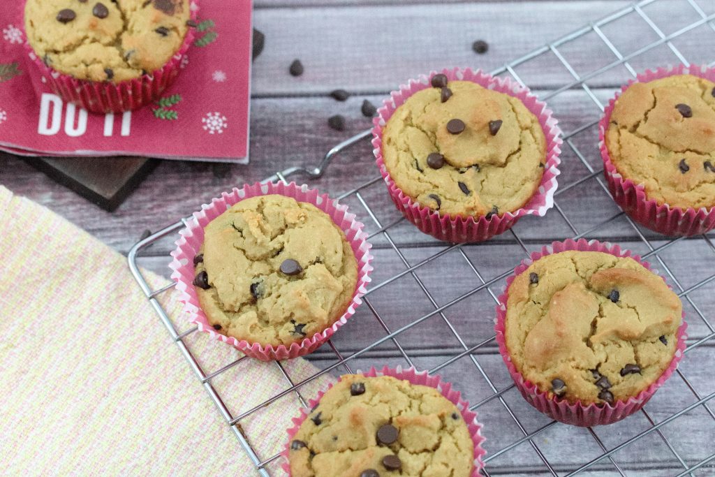 Gluten-free protein muffins with chocolate chips in red muffin cups on a baking tray with more chocolate chips behind them and another muffin on a red napkin