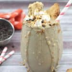 Glass with cookies and cream shake topped with coconut whipped cream and crumbled cookies with a striped straw in it. There are straws, chocolate chip, cookies and chocolate chips around the glass.