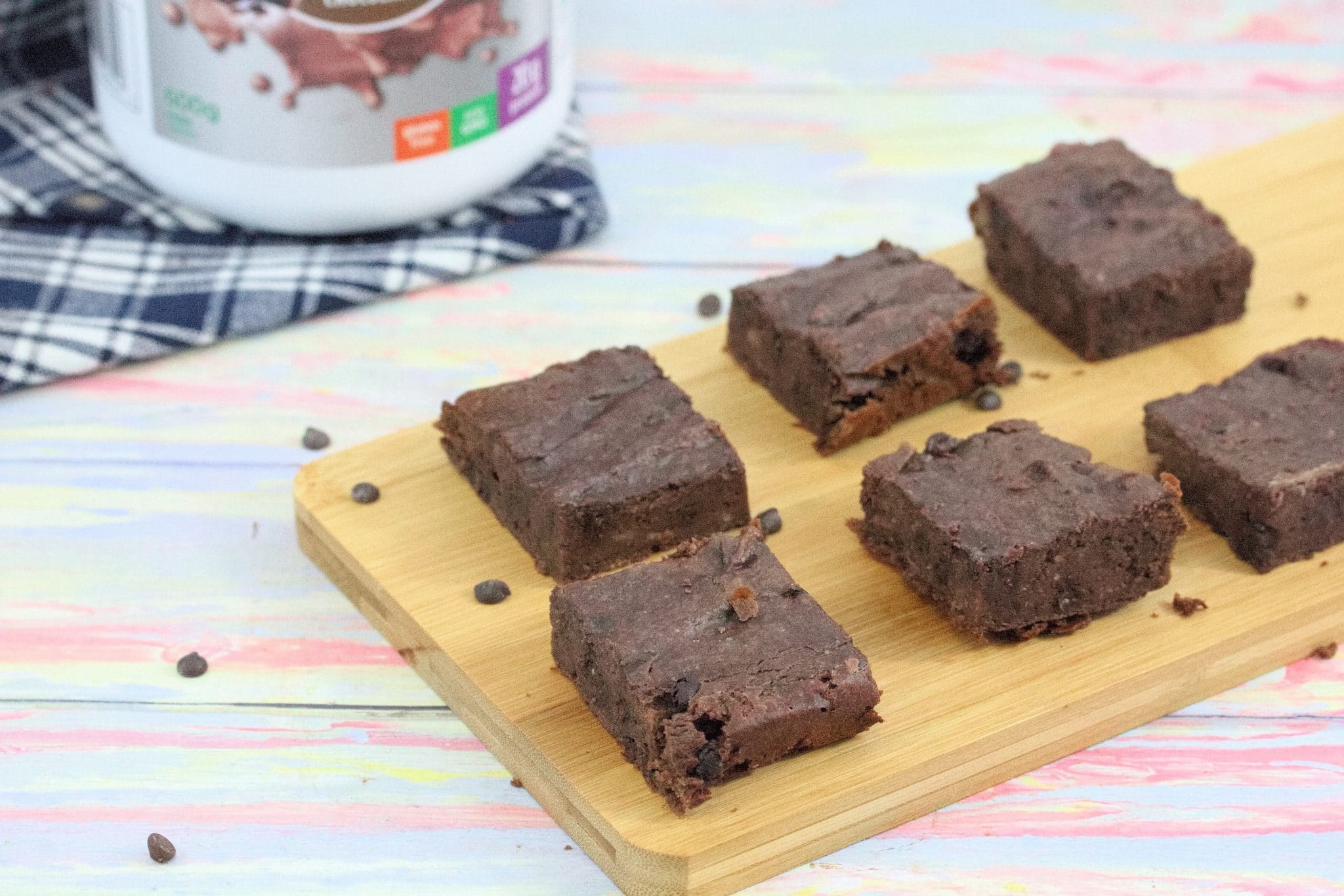 Six chocolate protein brownies on a wooden cutting board with chocolate chips around them and a jug of protein powder behind them