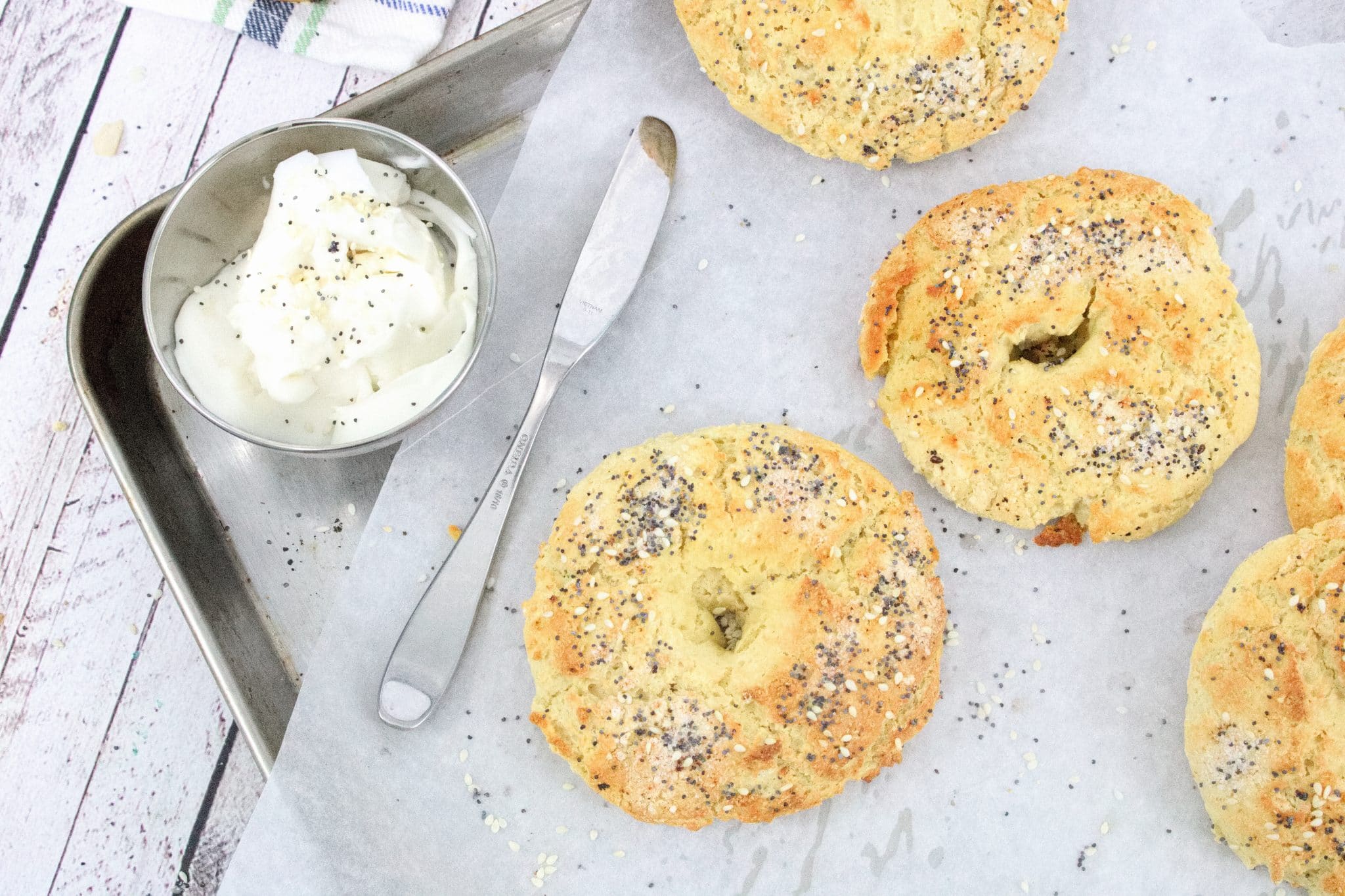multiple gluten-free bagels topped with everything seasoning on a baking sheet with parchment paper and cream cheese in a bowl beside it