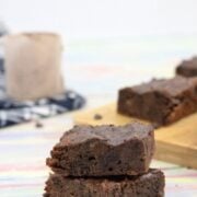 Two chocolate brownies stacked with more brownies on a cutting board and a scoop of protein powder on a blue linen