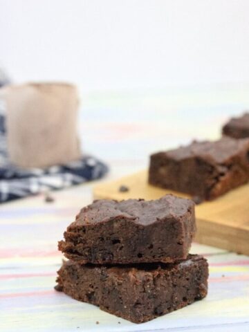Two chocolate brownies stacked with more brownies on a cutting board and a scoop of protein powder on a blue linen