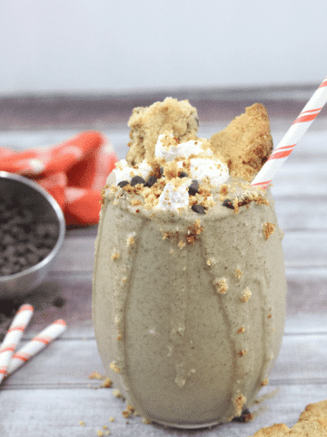 Glass with cookies and cream shake topped with coconut whipped cream and crumbled cookies with a striped straw in it. There are straws, chocolate chip, cookies and chocolate chips around the glass.