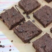 Dairy-free chocolate brownies drizzled with chocolate sauce on brown parchment paper with chocolate chips around them and a glass of milk with a blue linen to the side