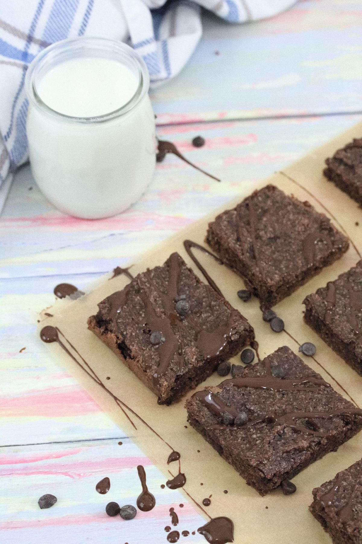 Dairy-free chocolate brownies drizzled with chocolate sauce on brown parchment paper with chocolate chips around them and a glass of milk with a blue linen to the side.
