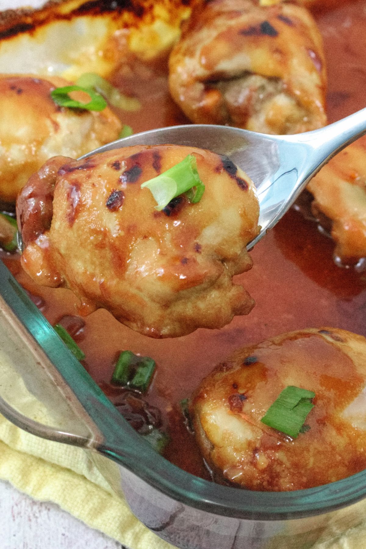 Cooked teriyaki chicken thighs in sauce in a glass baking tray with green onions on top and a spoon holding one chicken thigh up from the tray
