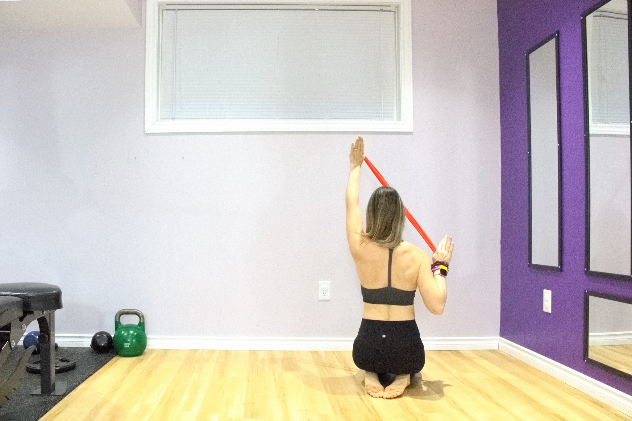 Woman in black sports bra and black pants sitting on her knees while pulling a red band down from overhead, back to camera