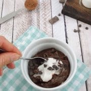 White ramekin on blue checkered linen with chocolate protein pudding and coconut cream in it and a spoon scooping some out with chocolate chips and collagen behind it