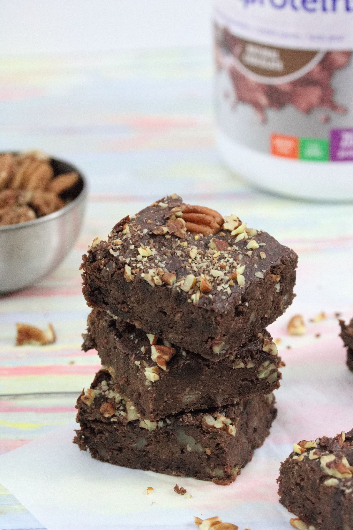 Three chocolate protein brownies stacked with protein powder behind them.