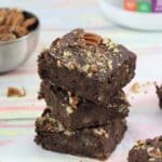 Three chocolate protein brownies stacked with protein powder behind them.