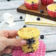 Three blueberry protein muffins with the top muffin being placed on top, all muffins on a pink muffin liner with more muffins on a cutting board and yellow linen behind them