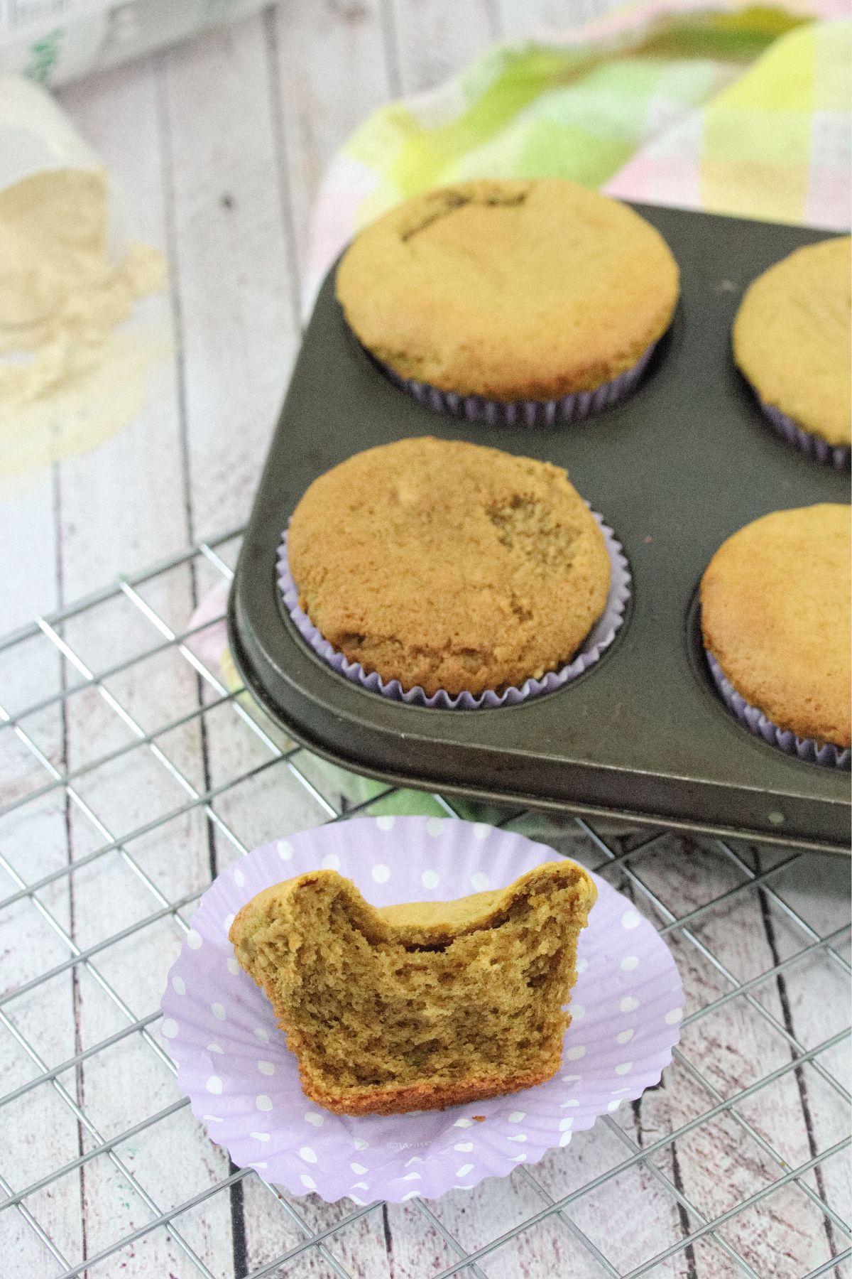 Half a protein muffin on a purple muffin cup on a cooling rack with more muffins in a muffin tray behind it.