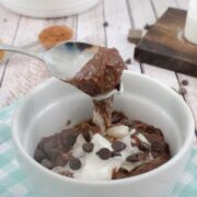 White ramekin on blue checkered linen with chocolate protein pudding and coconut cream in it and a spoon scooping some out with chocolate chips and collagen behind it.