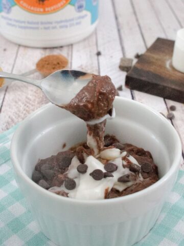 White ramekin on blue checkered linen with chocolate protein pudding and coconut cream in it and a spoon scooping some out with chocolate chips and collagen behind it.