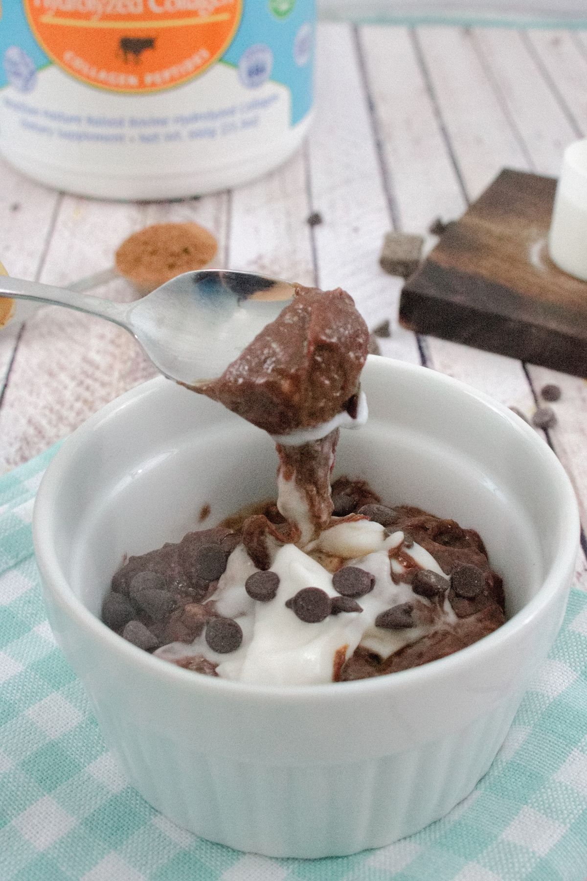 White ramekin on blue checkered linen with chocolate protein pudding and coconut cream in it and a spoon scooping some out with chocolate chips and collagen behind it.