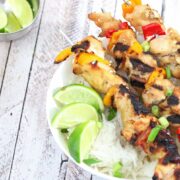 Three teriyaki chicken skewers on a bed of rice in a white bowl with limes on it and a bowl of limes beside it