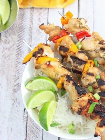 Three teriyaki chicken skewers on a bed of rice in a white bowl with limes on it and a bowl of limes beside it