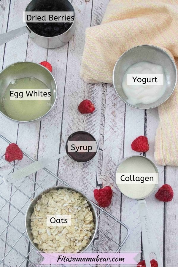 Collagen powder, rolled oats, egg whites, maple syrup, dried berries and baking powder in measuring dishes with labels and fresh raspberries around the bowls and an orange linen behind them