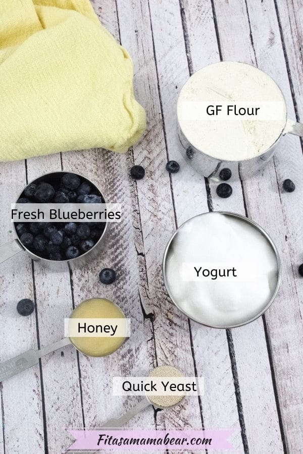 Multiple ingredients like yogurt, blueberries, honey, and flour in measuring cups with blueberries around them