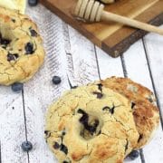A gluten-free blueberry bagel cut in half and stacked with another bagel on a yellow linen and more on a cutting board with a honey stick behind them