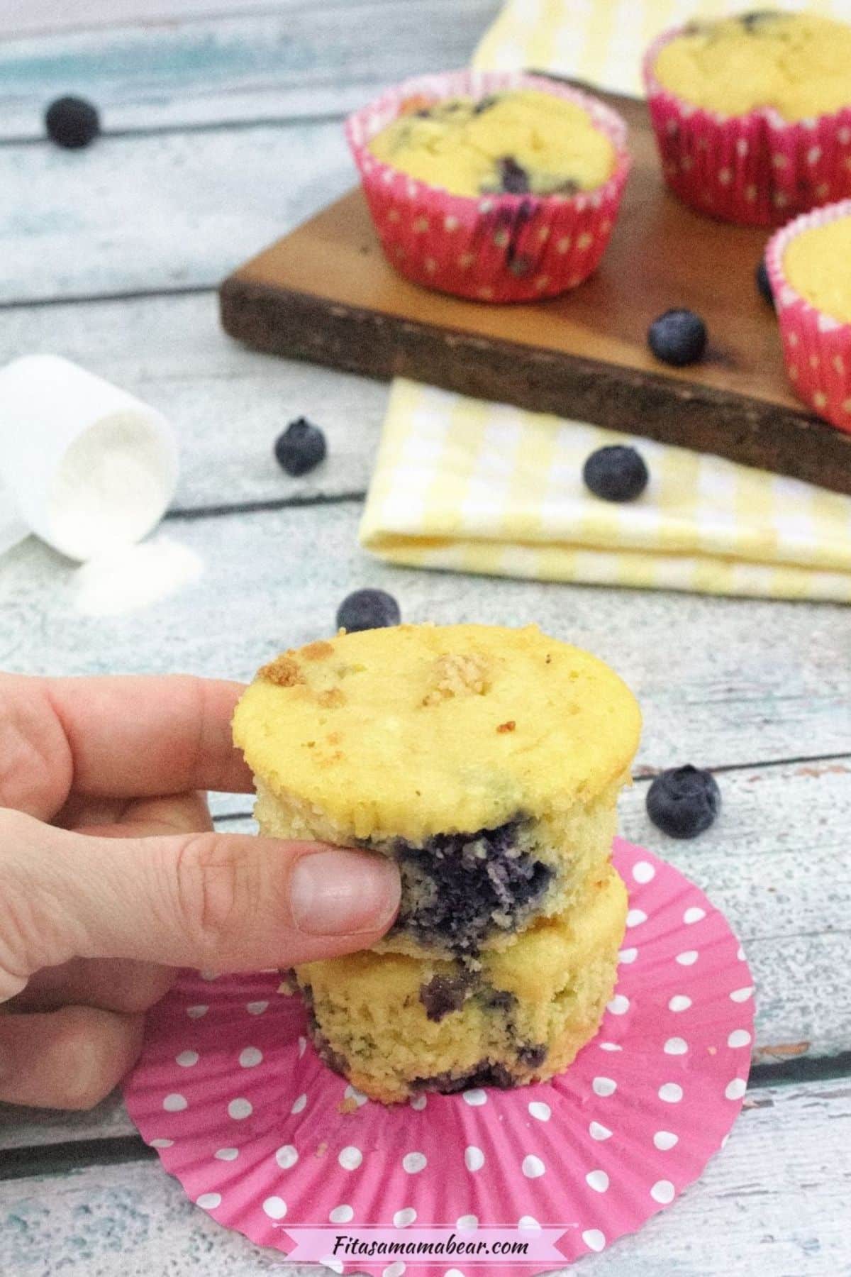 A person holding up a blueberry muffin on a pink paper plate.