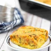 Egg casserole on a white plate with more casserole in a pan behind it and a blue linen with steel bowls on top beside it