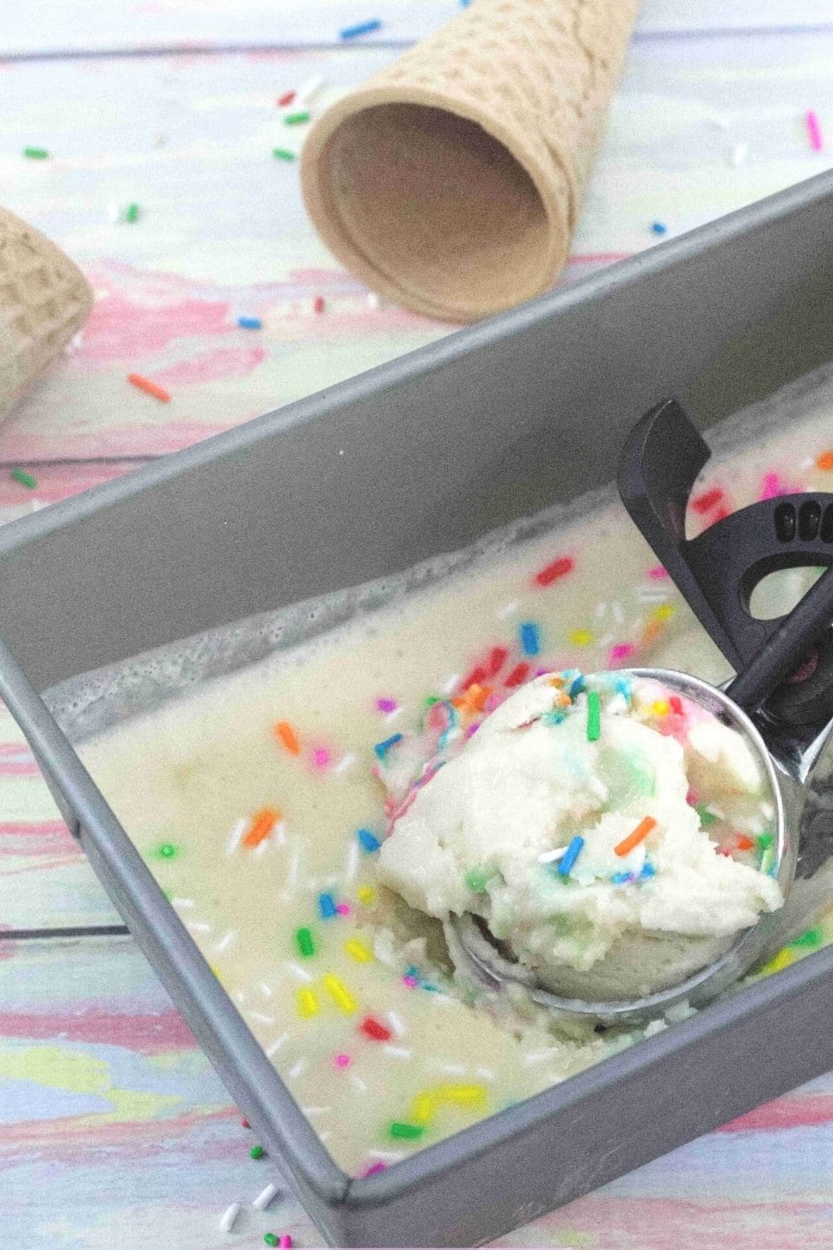 Black and steel ice cream scoop t scoop out vanilla protein ice cream with sprinkles from a baking tin with ice cream cones and sprinkles around it.