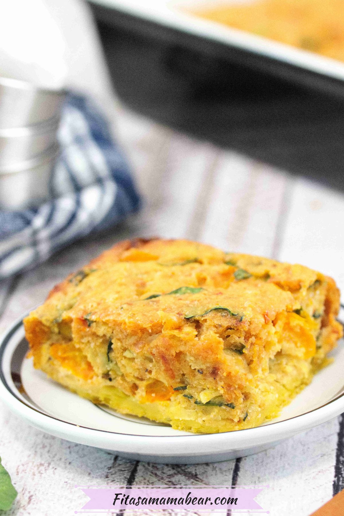 Egg casserole on a white plate with more casserole in a pan behind it and a blue linen with steel bowls on top beside it.