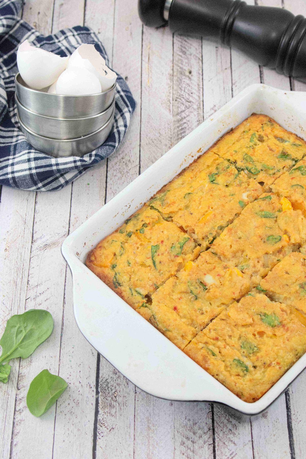 Dairy-free egg casserole baked in a pan with spinach around it and a blue linen with tin cups and egg shells behind it