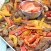 Baking tray with cooked steak, bell peppers, and veggies with a steel bowl of fresh tomatoes in the middle and flour tortillas folded to the side.