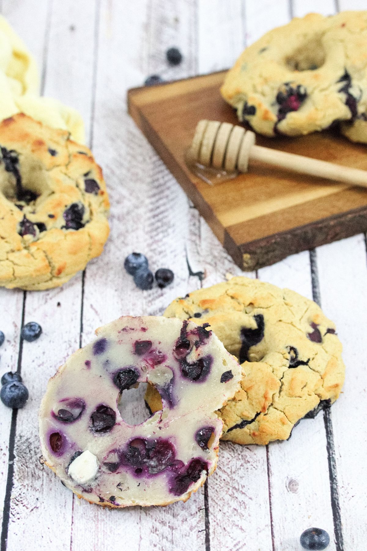 A blueberry bagel cut in half and openfaced smeared with butter with another bagel behind it and more on a dark cutting board with a stick of honey.