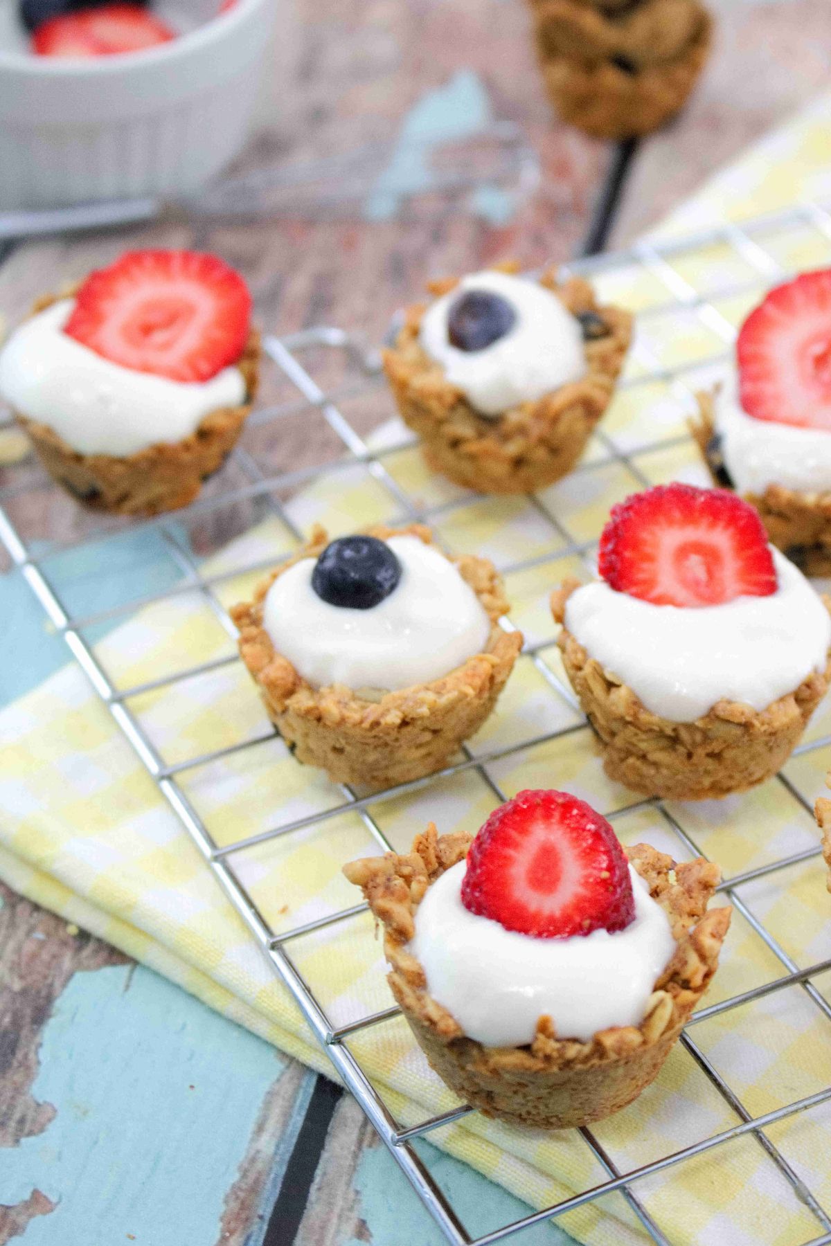 Mini granola cups topped with yogurt and blueberries and strawberries on a cooling rack over a yellow linen.