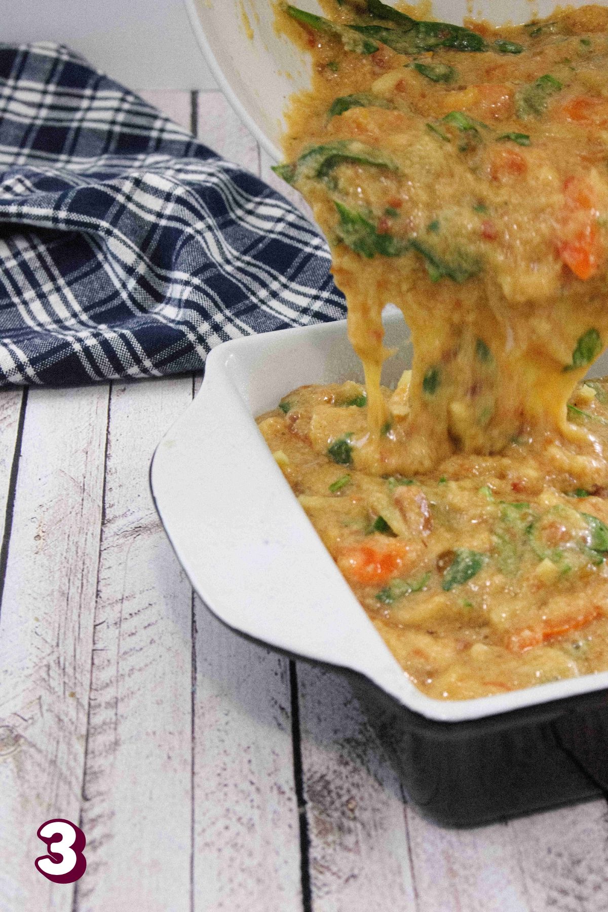 Raw breakfast egg casserole with peppers and spinach being poured into a white baking dish and blue linen behind it.