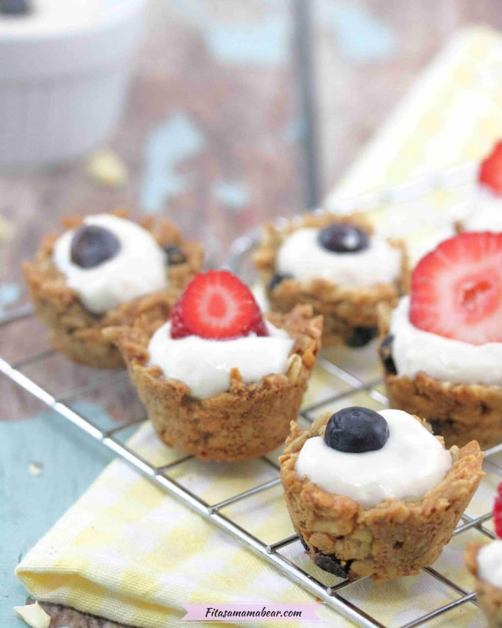 Mini granola cups topped with yogurt and blueberries and strawberries on a cooling rack over a yellow linen