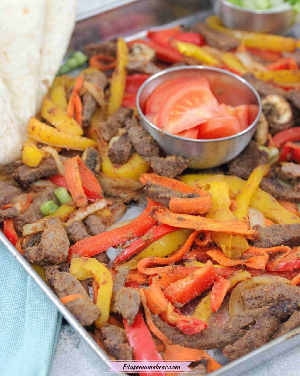 Baking tray with cooked steak, bell peppers, and veggies with a steel bowl of fresh tomatoes in the middle and flour tortillas folded to the side