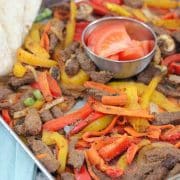 Baking tray with cooked steak, bell peppers, and veggies with a steel bowl of fresh tomatoes in the middle and flour tortillas folded to the side