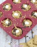 Pin image with text: Mini s'mores cups with graham cracker crust, chocolate, peanut butter and mini marshmallows in a mini, silicone red muffin tin on a blue linen