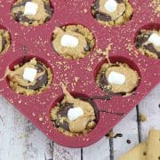 Pin image with text: Mini s'mores cups with graham cracker crust, chocolate, peanut butter and mini marshmallows in a mini, silicone red muffin tin on a blue linen