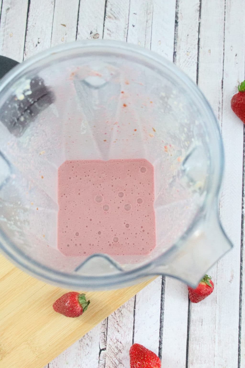 Strawberry protein shake in a blender on a cutting board with berries around it
