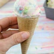 hand holding vanilla protien ice cream with sprinkles in a gluten-free ice cream cone with cones, sprinkles and the ice cream tin behind it