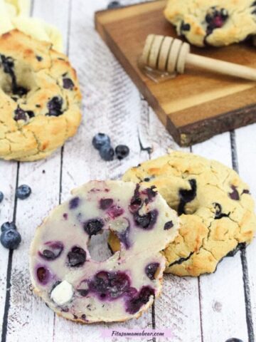 A blueberry bagel cut in half and openfaced smeared with butter with another bagel behind it and more on a dark cutting board with a stick of honey