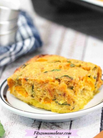 Egg casserole on a white plate with more casserole in a pan behind it and a blue linen with steel bowls on top beside it.