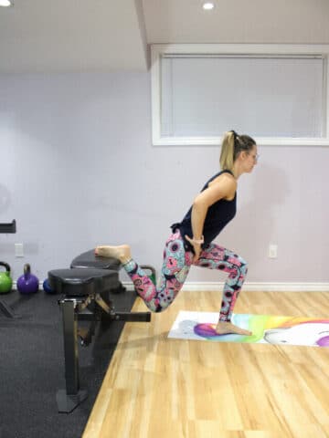 Woman in colored tights and a black tank top performing a bulgarian split squat with one foot on a workout bench