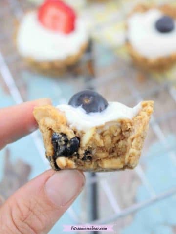 a close up image of two fingers holding a granola cup with yogurt and blueberry on top and the cup has been bitten in half