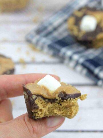Pin image with text: peanut butter s'mores cup with a marshmallow on top held between two fingers with more smores cups behind it