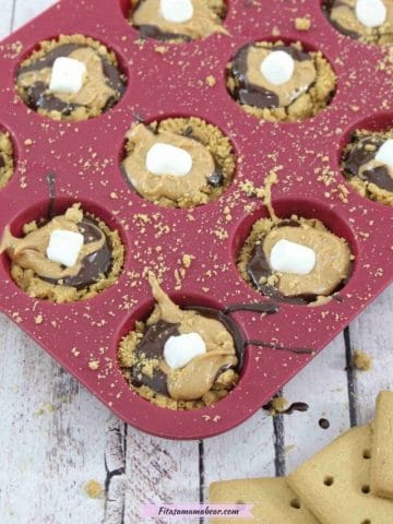 Pin image with text: Mini s'mores cups with graham cracker crust, chocolate, peanut butter and mini marshmallows in a mini, silicone red muffin tin on a blue linen