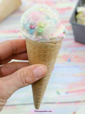 hand holding vanilla protien ice cream with sprinkles in a gluten-free ice cream cone with cones, sprinkles and the ice cream tin behind it