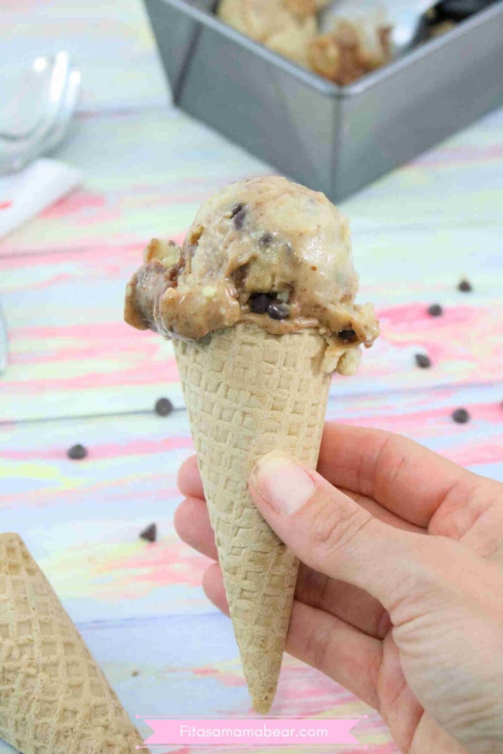 A chocolate swirled protein ice cream in an ice cream cone bring held up close with a baking load of protein ice cream behind it